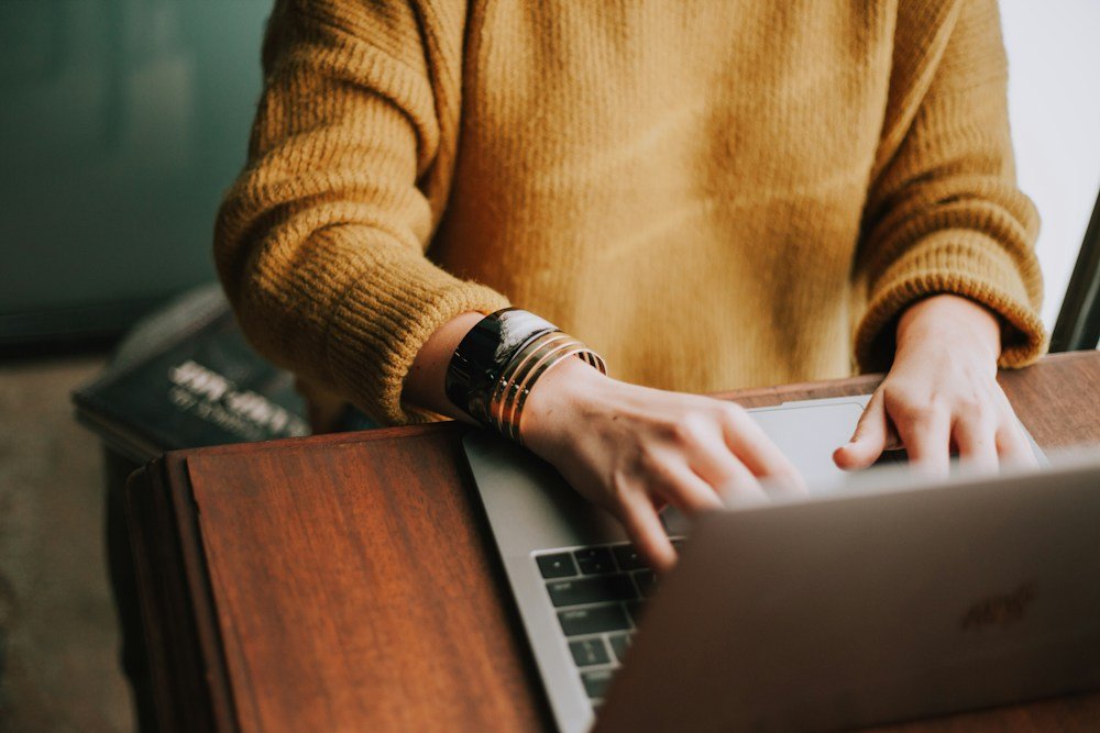 Person in a mustard jumper typing on a laptop at a wooden table